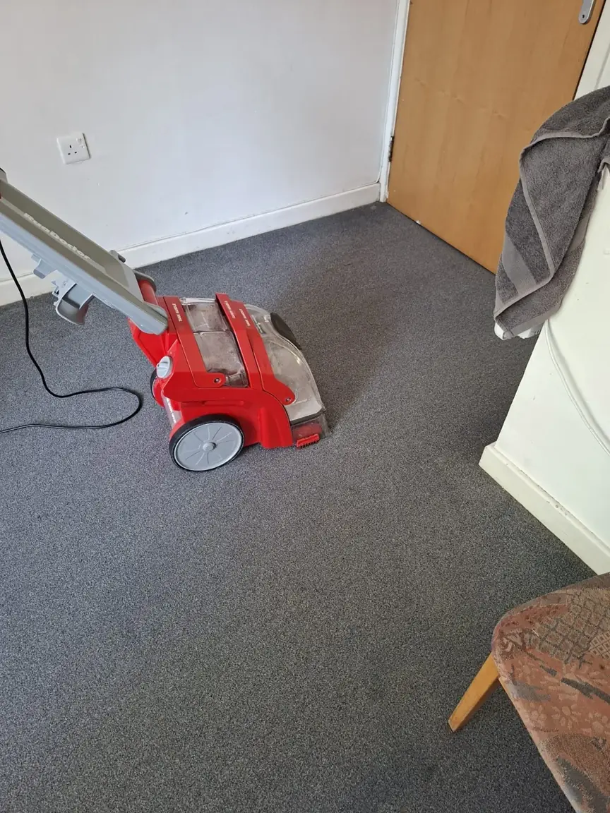 Red carpet-cleaning machine on grey carpet beside a white wall and wooden furniture